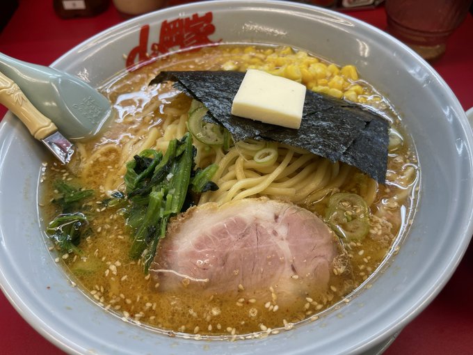 A bowl of Japanese ramen with chashu pork, nori, butter, corn, and green onions in a sesame-flecked broth, with a spoon resting on the side.