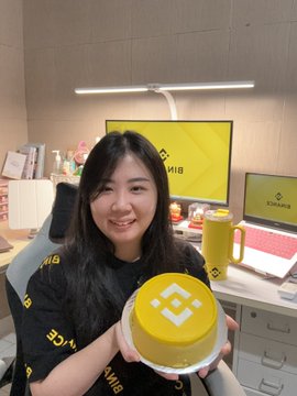 A woman wearing a black shirt with yellow letters is sitting at her desk holding up a small yellow cake decorated with a white "B" logo, which appears to be a birthday celebration for herself or someone else. The desk also has a laptop on it, indicating that she might have been working from home during the celebration.