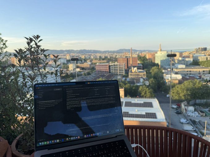 The image shows a laptop computer sitting on top of an outdoor table, with a view of a city skyline visible through the window. The laptop screen displays some text or code, suggesting that someone might be working from their rooftop location.