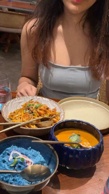 A photo of a woman at a restaurant wearing a light blue top, seated in front of several colorful dishes including blue rice, orange curry soup, and a stir-fry dish.
