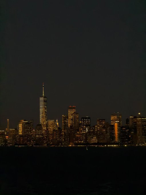 A nighttime photograph captures a city skyline illuminated against a dark sky, prominently featuring a tall skyscraper with a lit spire, likely the One World Trade Center.