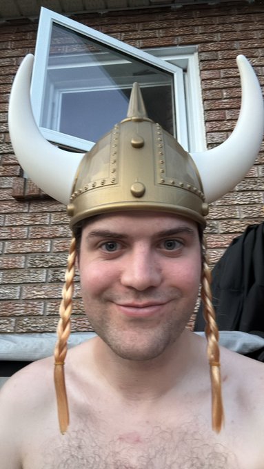 A shirtless man smiles while wearing a metallic gold Viking helmet with large white horns and braided ties, embodying the playful "Pirate with a Viking hat" persona described in the caption. The brick wall and window behind him suggest a casual backyard or patio setting for his fun photo shoot.