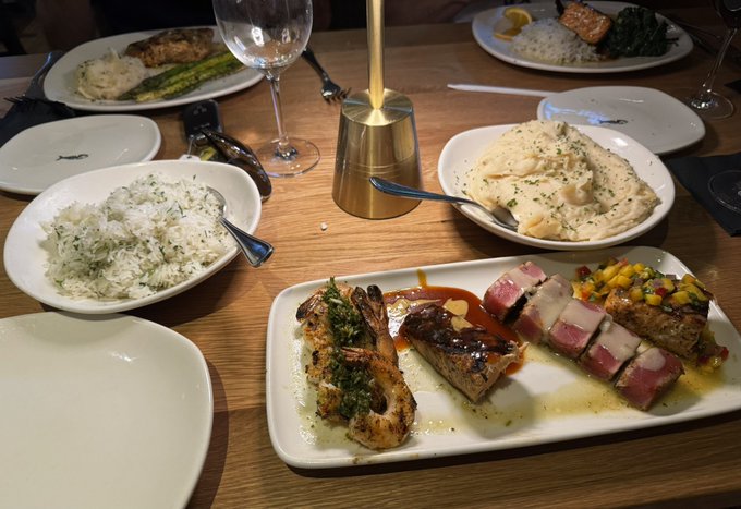 A wooden dining table set for a meal featuring several dishes, including a long platter with seared tuna, salmon, and shrimp, alongside bowls of rice and mashed potatoes.