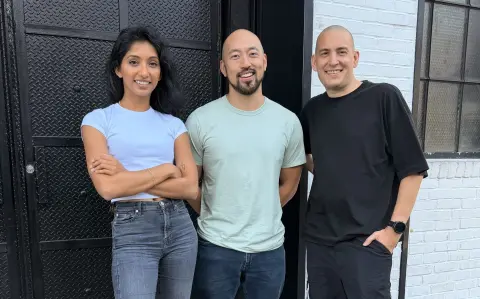 Three people pose together for a photo in front of a white brick building, with a woman in a blue shirt on the left, a man in a green shirt in the center, and a man in a black shirt on the right.
