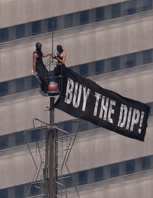 Two masked individuals on top of a communications tower unfurling a large black banner that reads "BUY THE DIP!" against a building facade.