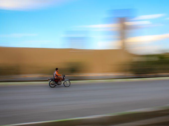 A motion-blurred photo of a person riding a motorcycle on a road against a backdrop of a tan wall and blue sky.