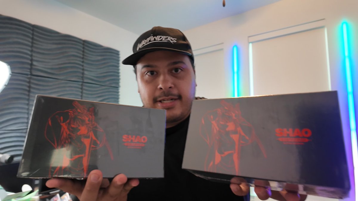 A man wearing a 'Wayfinders' cap holding two dark boxes with red anime-style artwork and the text 'SHAO' on them, in a room with neon blue lighting.