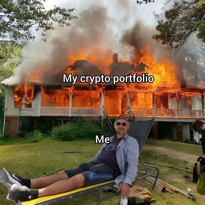 A meme image showing a man relaxing in a lounge chair in the foreground while his house burns down behind him. The text reads "My crypto portfolio" above the burning house and "Me" above the relaxed man, humorously depicting investor indifference to a market crash.