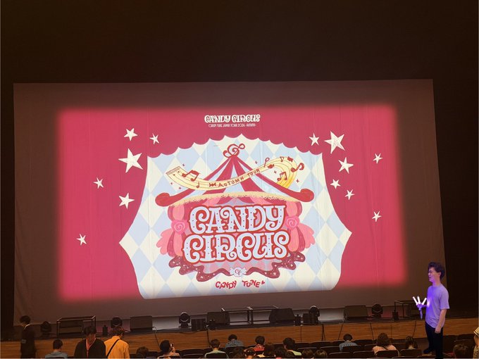 A presenter stands on stage next to a large screen displaying 'CANDY CIRCUS - A STRING OF CANDY -' with a circus tent logo, while audience members are seated in the foreground.
