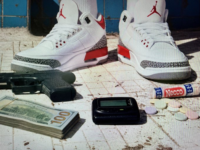 A stylized still life on a tiled surface featuring a person wearing white, red, and grey Air Jordan sneakers standing over a pile of items: a black handgun, a stack of $100 bills, a pager, scattered pills, and a Necco candy roll.