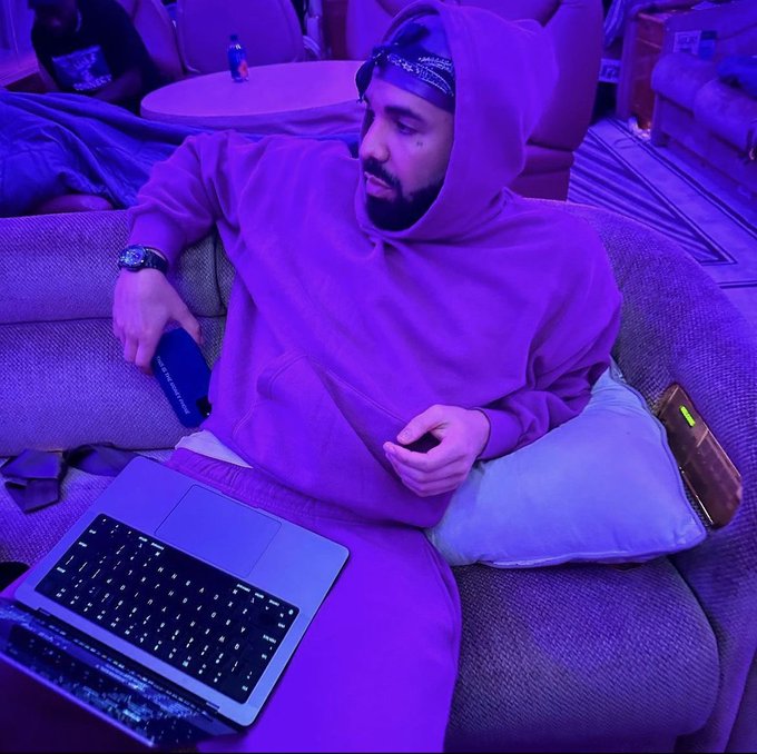 A man in a purple hoodie and matching sweatpants sits on a couch bathed in purple light, using a laptop while holding a gaming controller.