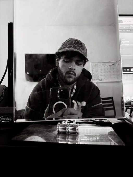 A black-and-white mirror selfie of a bearded man wearing a patterned cap, hoodie, and chain necklace, holding an iPhone with a MagSafe ring while sitting at a table with a salt shaker.