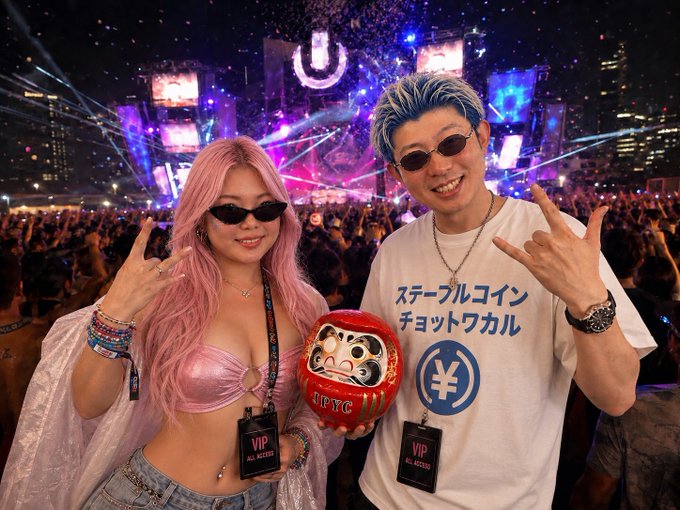 A woman with pink hair and a man in a white t-shirt with Japanese text pose with peace signs and a red Daruma doll labeled "UBYC" while wearing VIP "All Access" lanyards at the Ultra Music Festival. The Japanese text on his shirt translates roughly to "Stablecoin Chotto Wakuwaku," referencing cryptocurrency—a fitting visual for the note that the image is fabricated by AI.