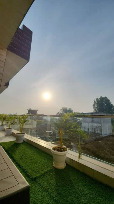 A rooftop balcony view at sunset or sunrise, featuring artificial green turf, potted plants, a glass railing, and a modern building structure on the left.