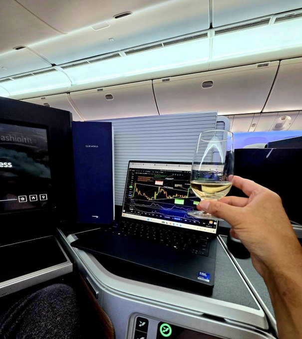 A hand toasts with a glass of champagne in an airplane cabin, with a laptop displaying a Bitcoin/cryptocurrency price chart featuring the BTC ticker visible on the screen. The first-class setting and celebratory toast align perfectly with the caption's mention of enjoying champagne while preparing to share monthly BTC and XRP charts on Patreon during the return flight home.
