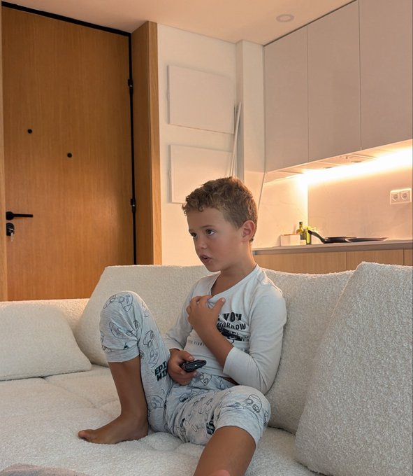 A young boy in pajamas sits on a couch holding a remote, looking focused. The caption celebrates his persistence, which reportedly helped his parent avoid a costly investment mistake—adding a humorous, heartfelt tone to the photo of the "annoying kid."