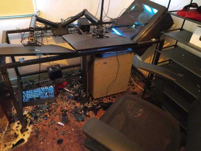 A trashed computer desk with a Dell monitor, mechanical keyboard, and broken chair sits amid scattered debris, paired with an angry post raging against "banking cartels," "greedy politicians," and "clarity delays." The chaotic workspace visually mirrors the poster's frustrated rant, suggesting the destroyed setup is a symbolic protest against their grievances.
