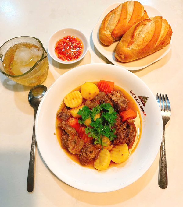 A meal featuring braised beef with potatoes and carrots, served with crusty bread, chili sauce, and a beverage in a glass, with the bowl bearing a 'DAT XANH GROUP' logo.