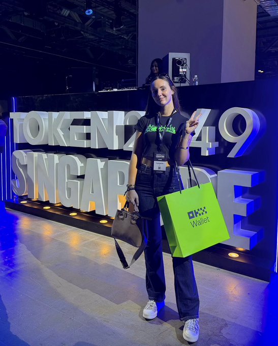 A woman wearing an OKX-branded shirt poses with a peace sign at a Token2049 Singapore event, holding an OKX Wallet shopping bag.