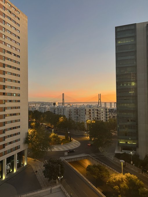 The image captures a sunset view over an urban landscape with modern buildings in the foreground and a bridge silhouetted in the distance, embodying a fresh and uplifting start to the week. The warm golden hues reflect an optimistic "Good Morning and Happy New Week" sentiment.