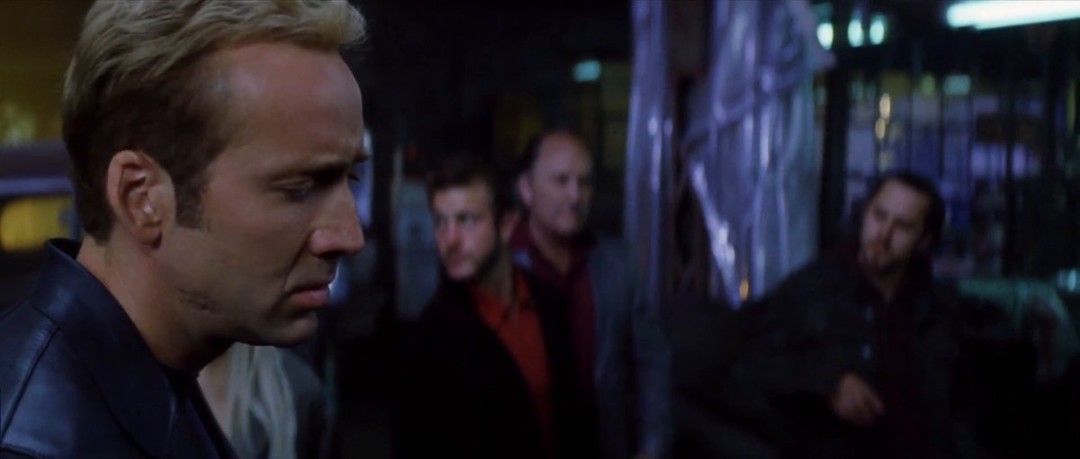 The image shows four men standing together, with one man looking down at his phone while the others look up towards him. The scene appears to be set in an urban environment, possibly a bar or a restaurant, as indicated by the presence of a dining table and chairs nearby. One of the men is wearing a black jacket, which stands out against the dark background.