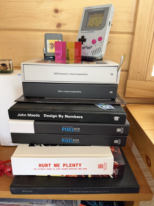A stack of design books including 'NES/Famicom: a visual compendium', 'N64: a visual compendium', 'Design By Numbers' by John Maeda, 'Pixel Book', and 'Hurt Me Plenty', topped with a Game Boy and cartridges.