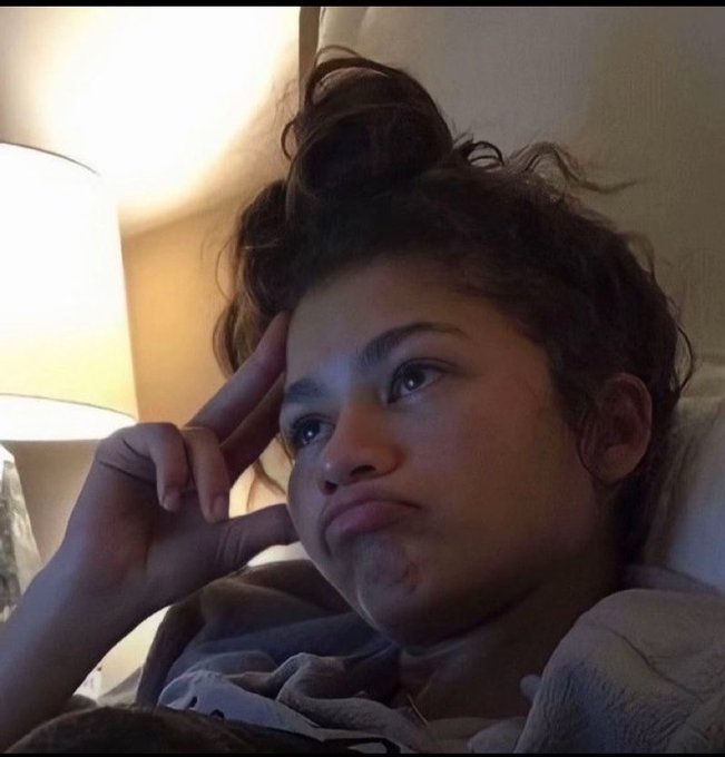 A selfie of a young woman with curly hair tied up, resting her head on her hand with a pouting expression, illuminated by warm light from a nearby lamp.