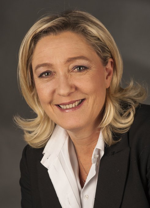 A professional headshot of a smiling woman with blonde hair wearing a white collared shirt and black blazer against a gray background.