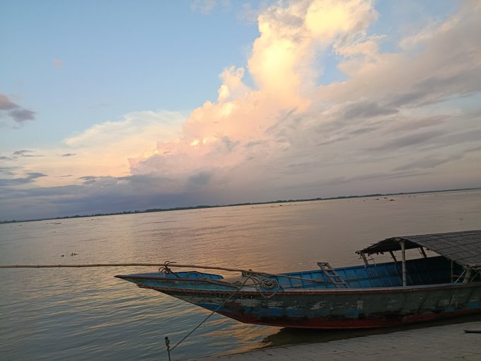 A serene landscape photograph of a weathered blue and red wooden boat moored near the shore of a calm body of water beneath a sky filled with clouds illuminated by the setting sun.