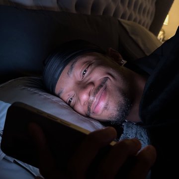 A smiling man wearing a black durag lies in bed in low light, holding a smartphone in his hand.