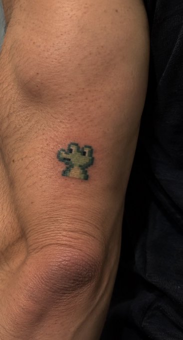 A small pixel-art style tattoo of a green creature, resembling a Minecraft design, is inked on a person's wrist.