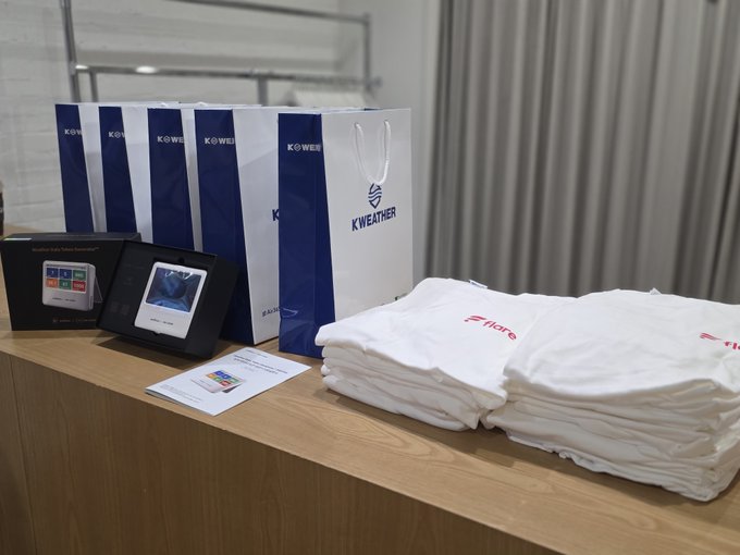 A product display featuring "K-WEATHER" branded shopping bags, a digital weather device box, a small monitor, an instruction manual, and two stacks of white Zippo-branded towels on a wooden surface.