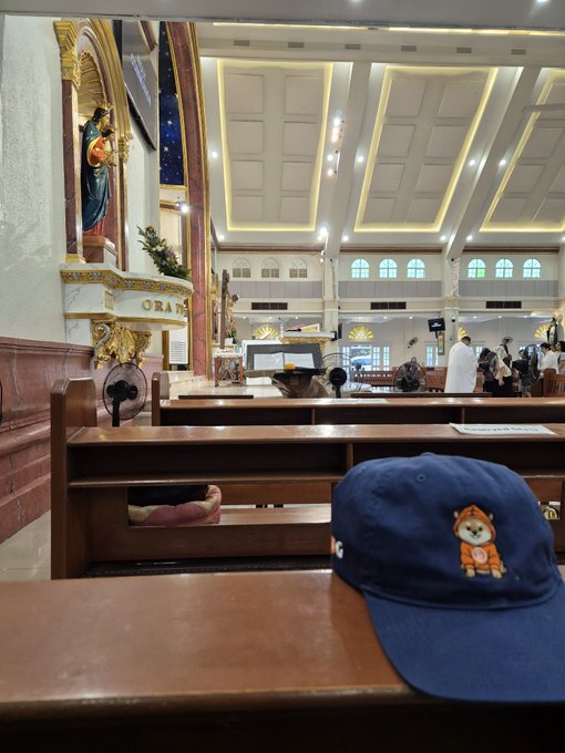 Photo of a blue cap with a Shiba Inu patch resting on a church pew, with the church's altar, statue, and ceiling visible in the background.