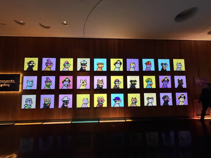 A dimly lit gallery space features a wall display of colorful pixel-art portraits arranged in a grid, showcasing various character designs against pastel backgrounds.
