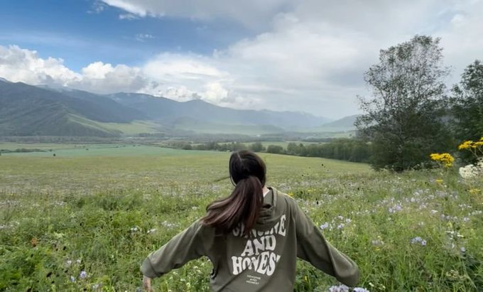 A person with long dark hair in a ponytail stands in a vast green meadow filled with wildflowers, wearing an olive green sweatshirt reading "SUNSHINE AND HOVES," looking out toward distant misty mountains.