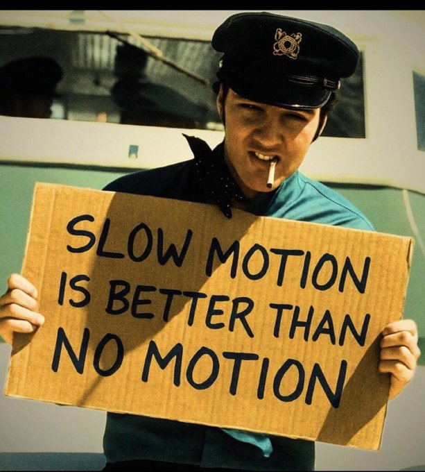 A person in a conductor's hat with a cigarette in their mouth holds a cardboard sign reading 'SLOW MOTION IS BETTER THAN NO MOTION'.