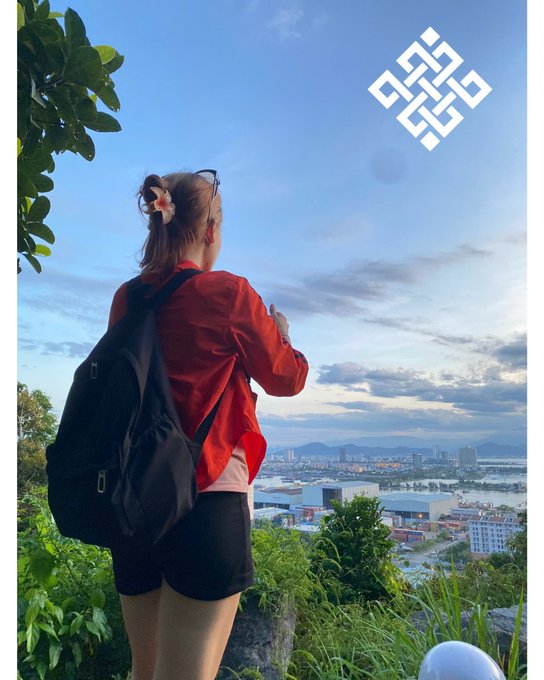A woman with a backpack, red jacket, and hair scrunchie standing on a hilltop overlooking a coastal city at sunset, with a decorative geometric logo in the upper right corner.