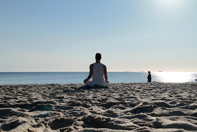 A man sits in a meditative pose on a sandy beach, gazing out at the calm ocean with another figure visible in the distance. The serene setting perfectly embodies the "Calmwave protocol" — a moment of mindfulness, stillness, and mental reset by the sea.
