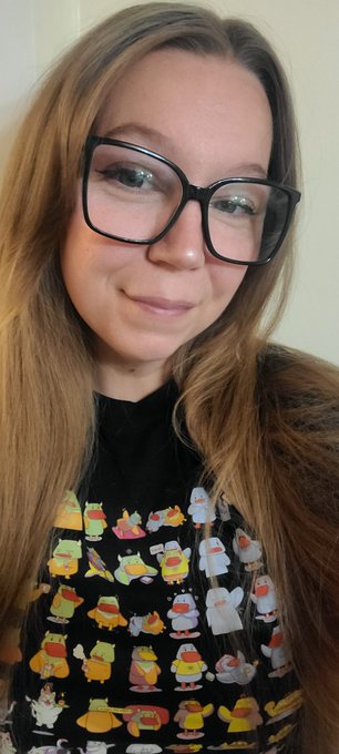 A young woman with long brown hair and black glasses wears a black shirt featuring a colorful print of cartoon characters.