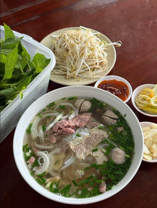 A bowl of Vietnamese pho with sliced beef and meatballs, accompanied by bean sprouts, fresh herbs, chili sauce, lemon, and garlic on a wooden table.