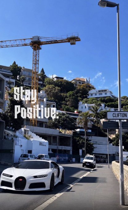 A scenic street view in Clifton featuring a white Bugatti Veyron sports car in the foreground, with a yellow construction crane, hillside houses, and the text 'Stay Positive.' overlaid.