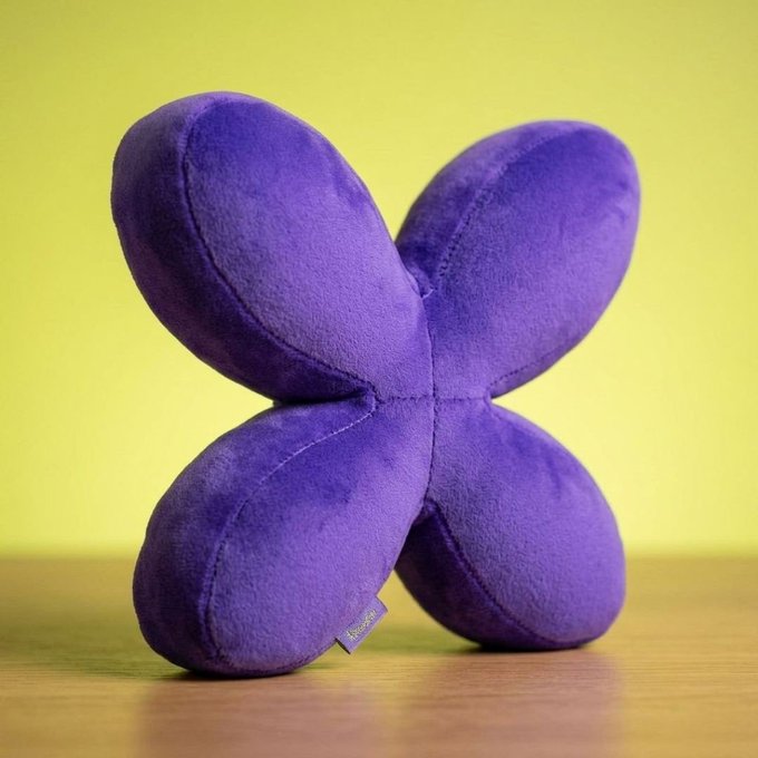 A purple plush Gflap—a squishy, X-shaped toy made of four rounded, pillowy segments connected at the center—sits on a wooden surface against a bright yellow background, with a small branded tag visible on the lower left lobe.