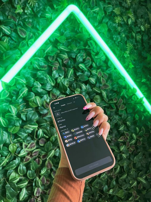 A hand holding a smartphone displaying a webpage for the crypto token 'StTicker' with various exchange logos, set against a green leafy wall background with a bright green neon triangle light.