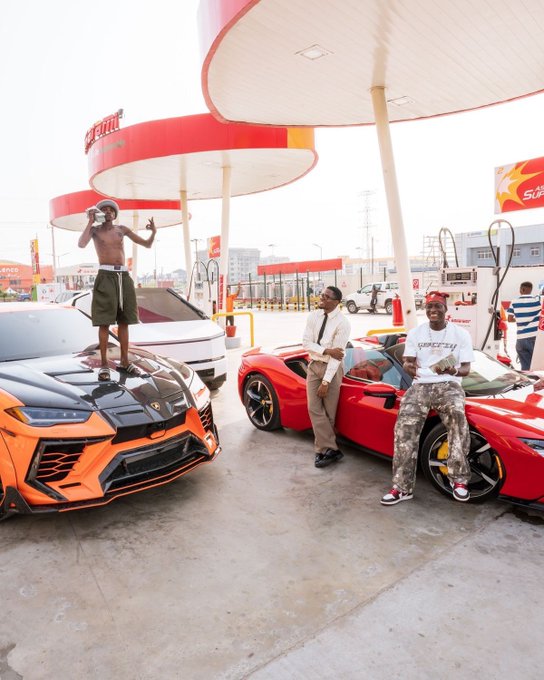Three men posing with luxury sports cars (an orange Lamborghini and a red Ferrari) at a gas station, with one shirtless man standing on the Lamborghini's hood.
