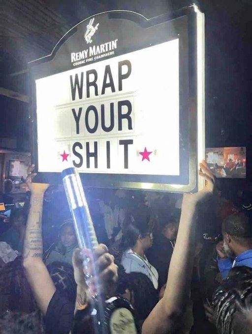A person in a crowded club holds up an illuminated Rémy Martin sign that reads 'WRAP YOUR SH*T' in bold letters.