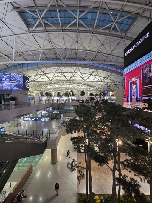 Interior view of a large, modern airport terminal with a geometric glass ceiling and an advertisement for 'CUBE MAGAZINE' on a digital display.