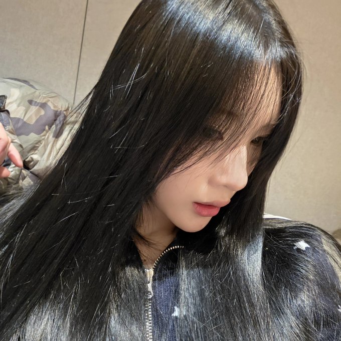 A close-up selfie of a young woman with long black hair and bangs, wearing a black fuzzy zip-up jacket with white star patterns, looking downward at the camera.