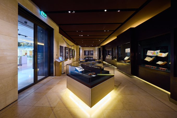A dimly lit, modern interior hallway featuring display cases under spotlights, with informational panels on the walls and an illuminated glass door.