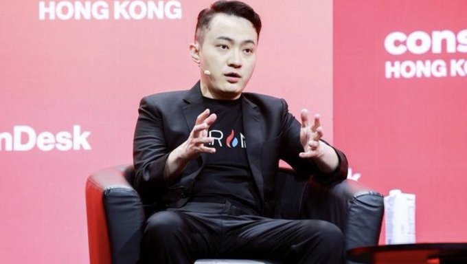 A man wearing a black blazer over a black t-shirt with a logo speaks while gesturing with both hands at an event with "HONG KONG" and "Consensus" branding visible on a red background.