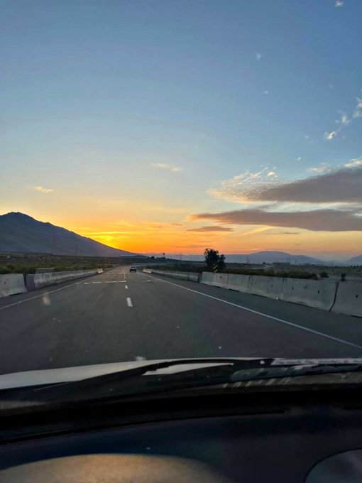 A sunset view from inside a vehicle on a multi-lane highway, with orange and yellow hues in the sky, mountains in the distance, and concrete barriers along the road.